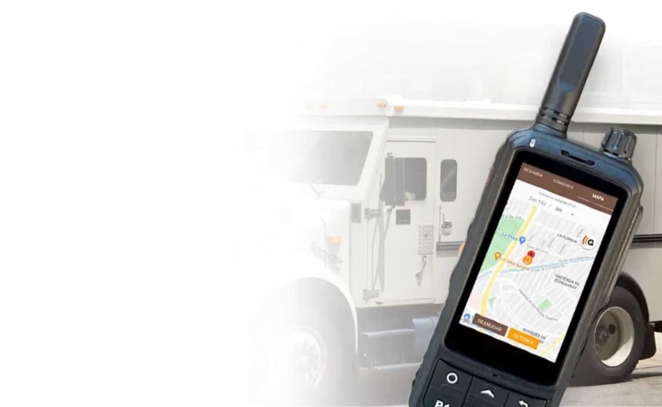 Un radio comunicador de uso rudo con pantalla que muestra un mapa de monitoreo GPS, en primer plano, con un camión blindado de traslado de valores en el fondo.