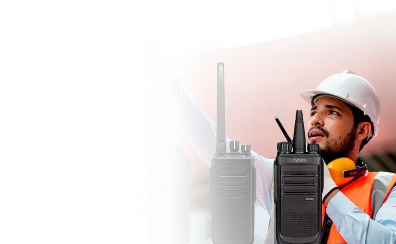 Ingeniero en una zona de carga, señalando un conjunto de radios de uso rudo de diferentes marcas con certificaciones de resistencia, con contenedores de carga de fondo.