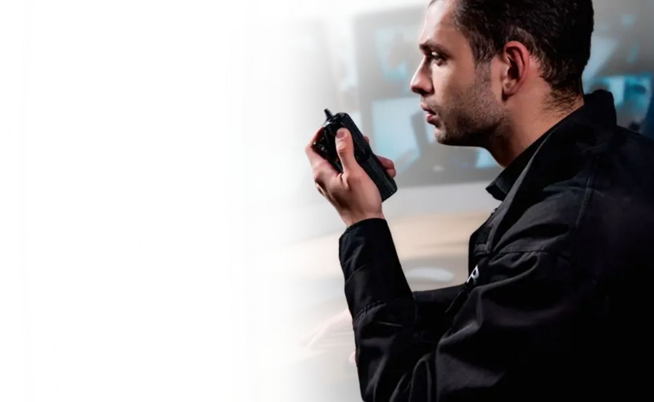 Guardia de seguridad privada en un centro de control, usando un radio de uso rudo y monitoreando cámaras de vigilancia de un vehículo en múltiples pantallas.