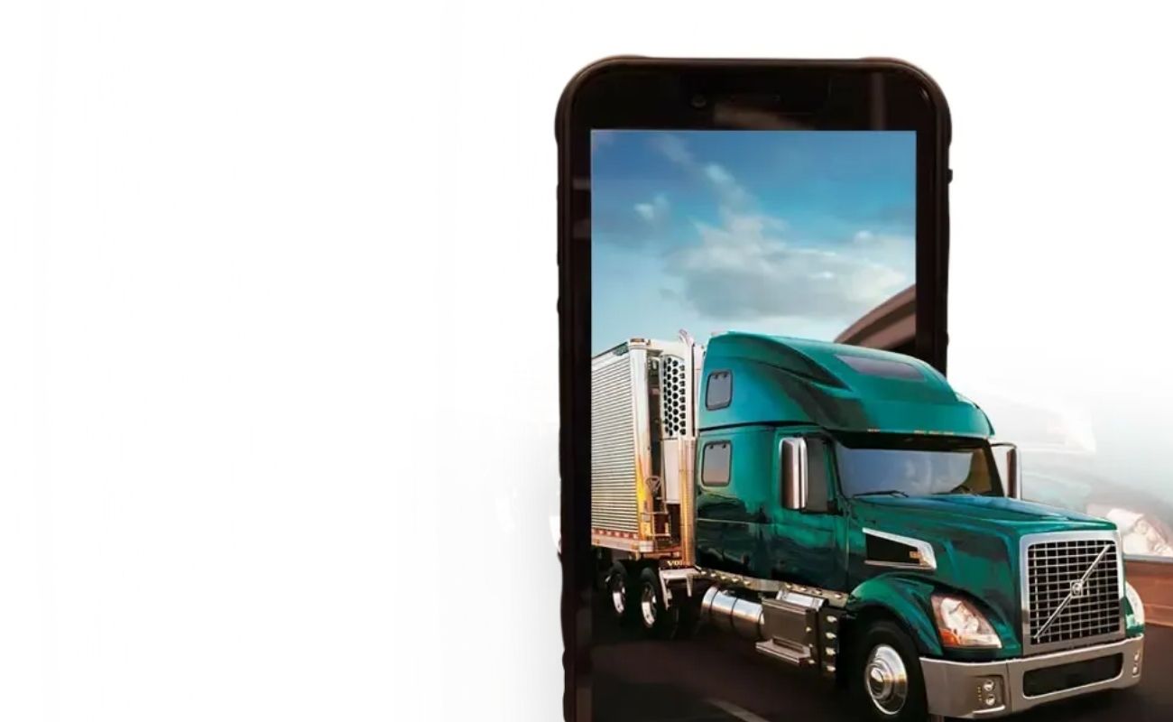 Un radio comunicador de uso rudo con pantalla que muestra un mapa de monitoreo GPS, en primer plano, con un camión de transporte de carga en el fondo.