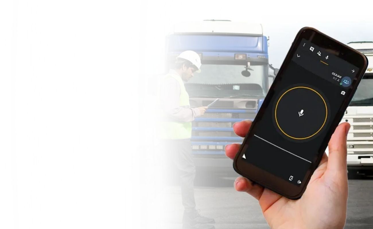 Mano sosteniendo un smartphone con una aplicación de radiocomunicación PTT, con un operador de autotransporte revisando un tablet con un camión de carga de fondo.