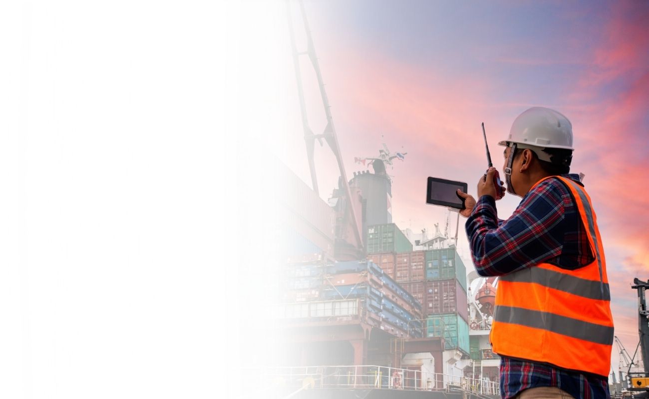 Operador portuario usando un radio de comunicación PTT y un tablet en un muelle de carga con un barco y contenedores de fondo.