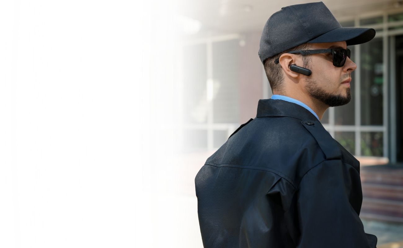 Guardia de seguridad privado con uniforme negro y auricular bluetooth, realizando vigilancia en la entrada de un edificio moderno.