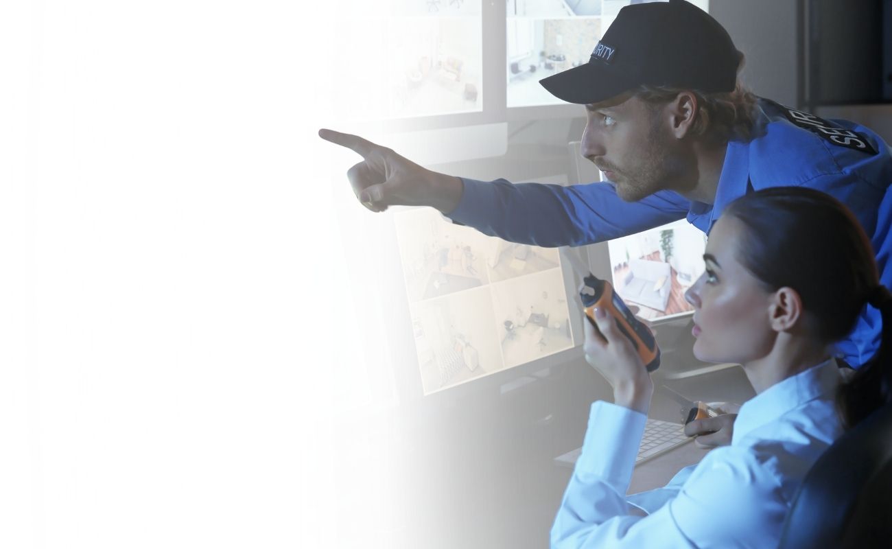 Dos operadores de seguridad privada en un centro de control, usando un radio PTT y monitoreando cámaras de videovigilancia en múltiples pantallas.