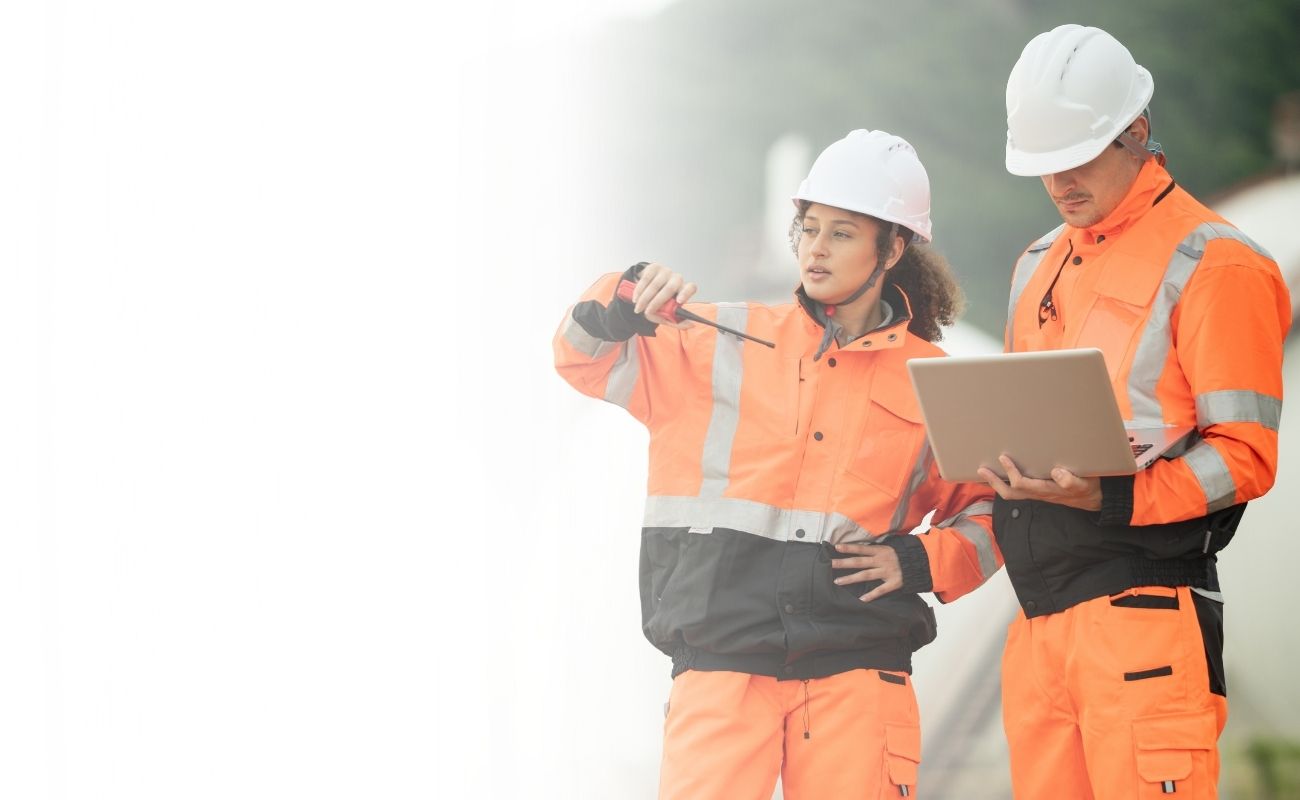 Mujer supervisora usando un radio de comunicación PTT mientras un ingeniero masculino revisa datos en una laptop, en una obra de infraestructura energética.