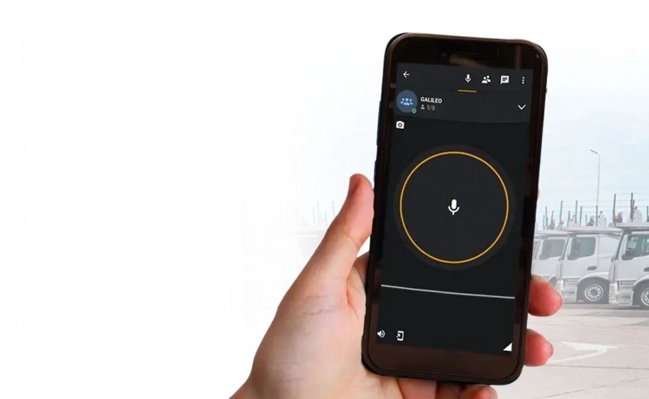 Mano sosteniendo un radio de uso rudo con la pantalla encendida mostrando la aplicación de comunicación PTT, con una flota de camiones de carga de fondo.