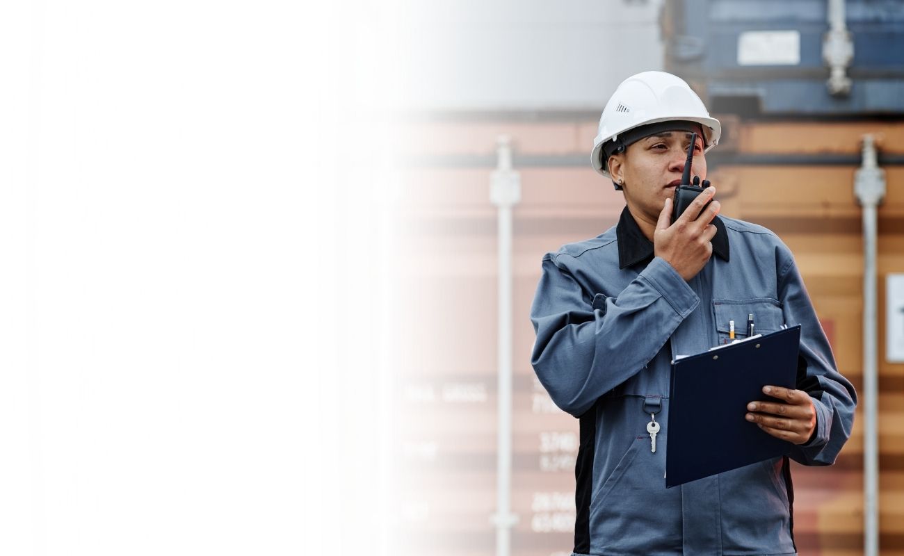Operaria de puerto o logística con casco de seguridad blanco, usando un radio PTT para coordinar el movimiento de contenedores de carga en un muelle.