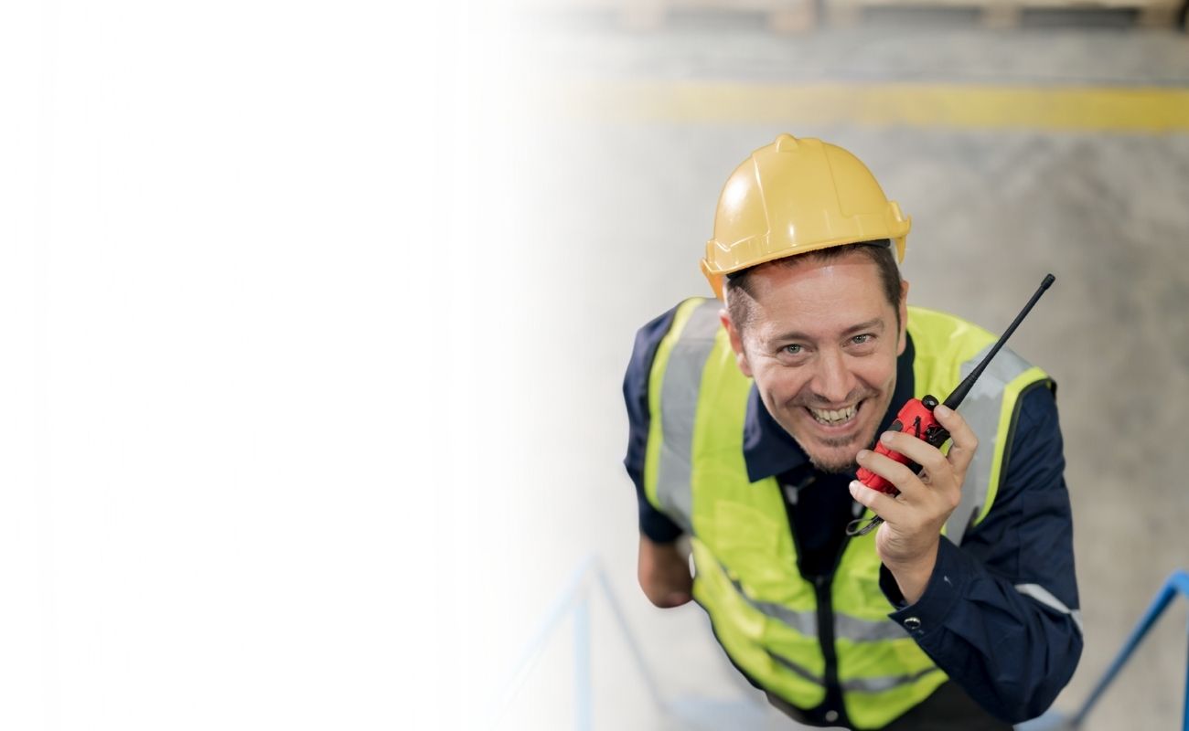 Operario de almacén o supervisor sonriendo, usando un radio de comunicación PTT en su mano para coordinar tareas en un almacén.