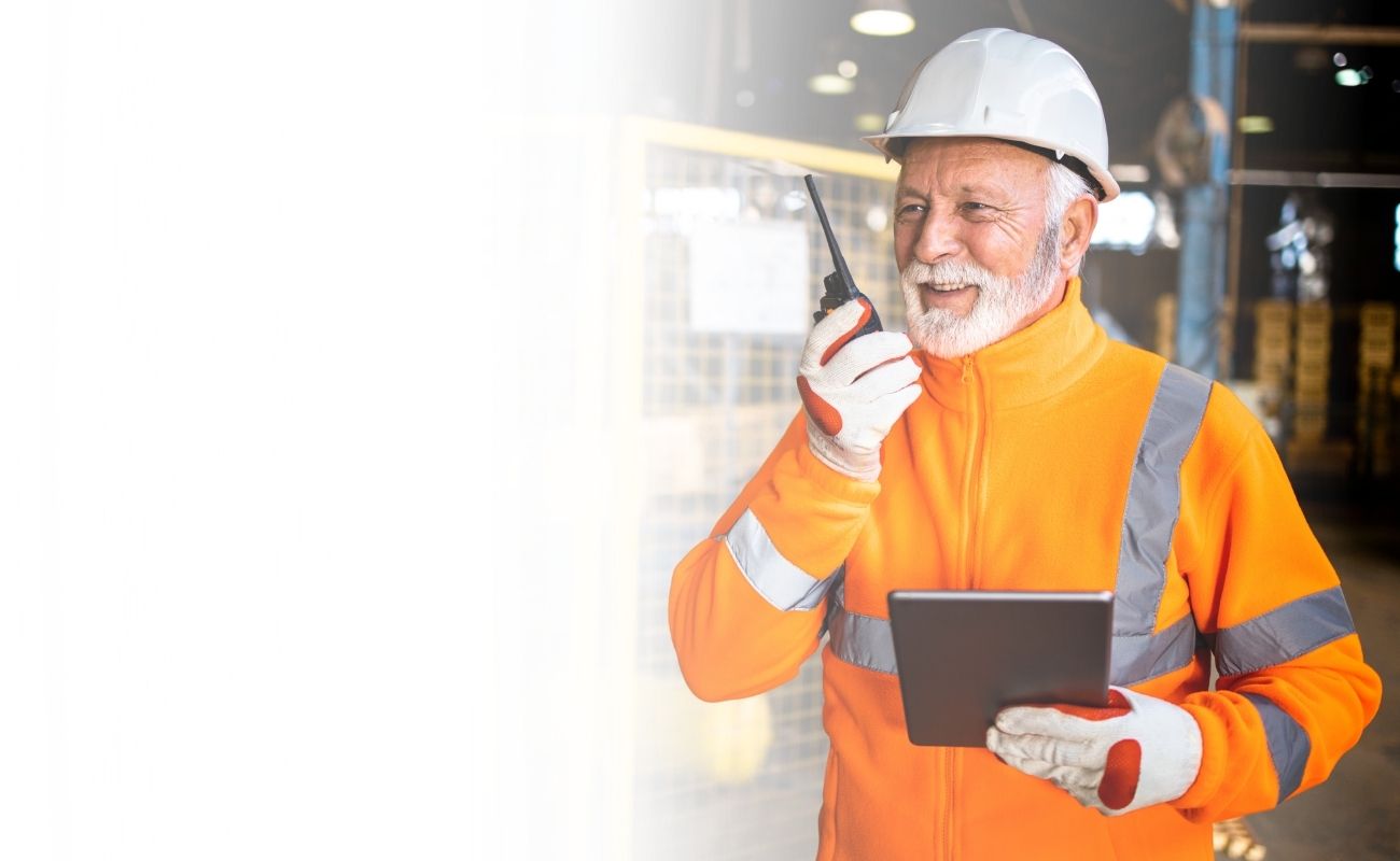 Operario industrial mayor, sonriendo y usando un radio de comunicación PTT y un tablet en el piso de una fábrica moderna.