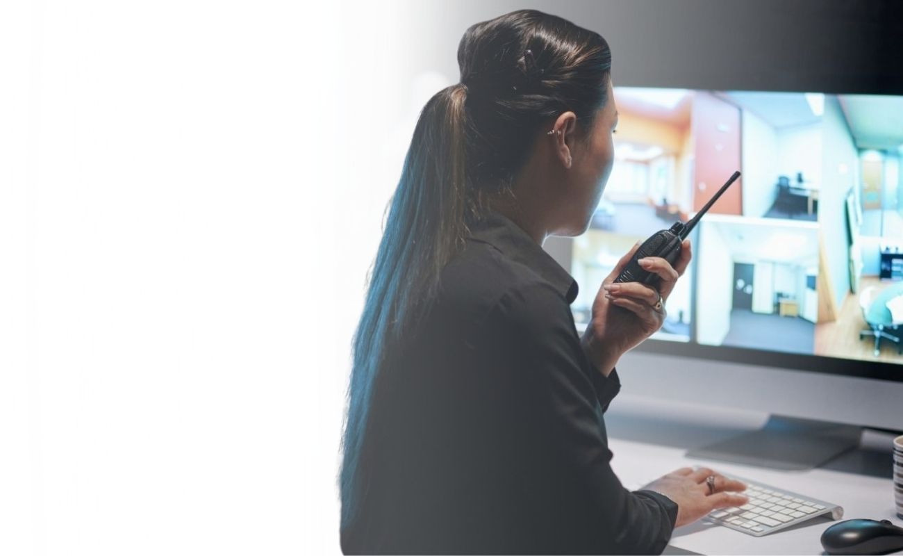 Operadora de seguridad monitoreando múltiples pantallas de videovigilancia y comunicación, con un radio PTT en mano, en un centro de control oscuro.