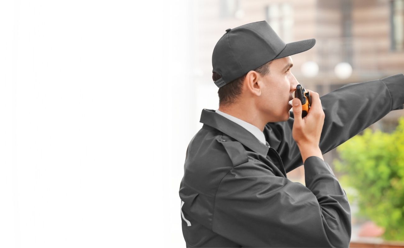 Guardia de seguridad privado con gorra y uniforme oscuro, usando un radio de comunicación PTT y señalando un punto mientras realiza vigilancia en la calle.