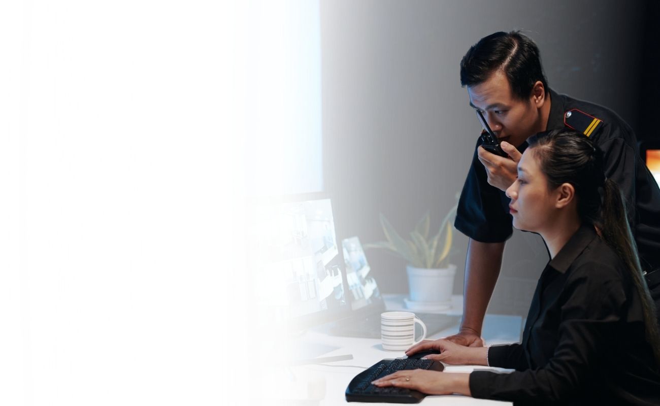 Operador de seguridad privada usando un radio de comunicación PTT, mientras es asistido por una colega que monitorea cámaras de vigilancia en múltiples pantallas.